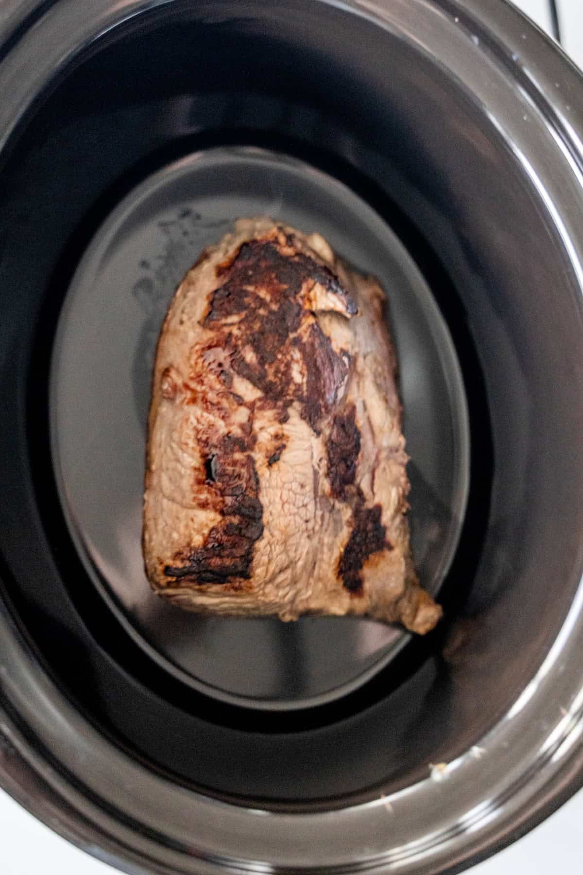 A seared piece of Slow Cooker Sirloin Steak rests inside the bottom of a black slow cooker, ready to be transformed into a tender and flavorful meal.