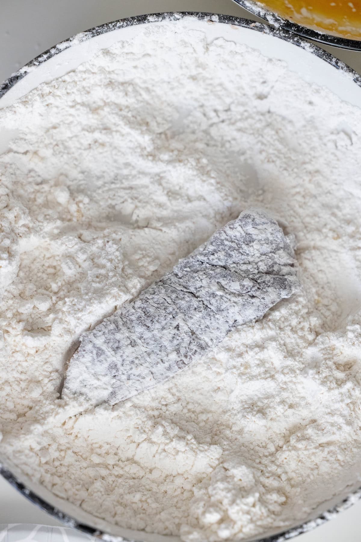A piece of fish coated in flour lies in a bowl filled with flour, ready for breading or frying—just like preparing steak fingers or a classic fried steak.