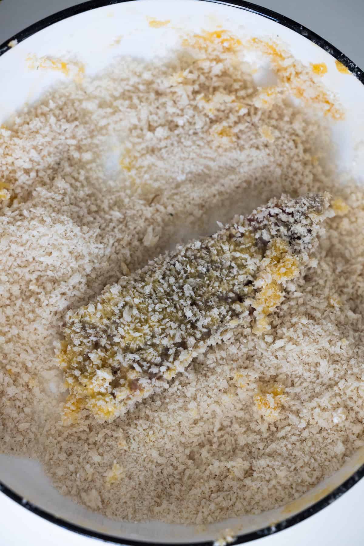 A piece of food, perfect for a Steak Fingers Recipe, is coated in egg and breadcrumbs and sits in a white bowl filled with more breadcrumbs, ready for frying or baking.