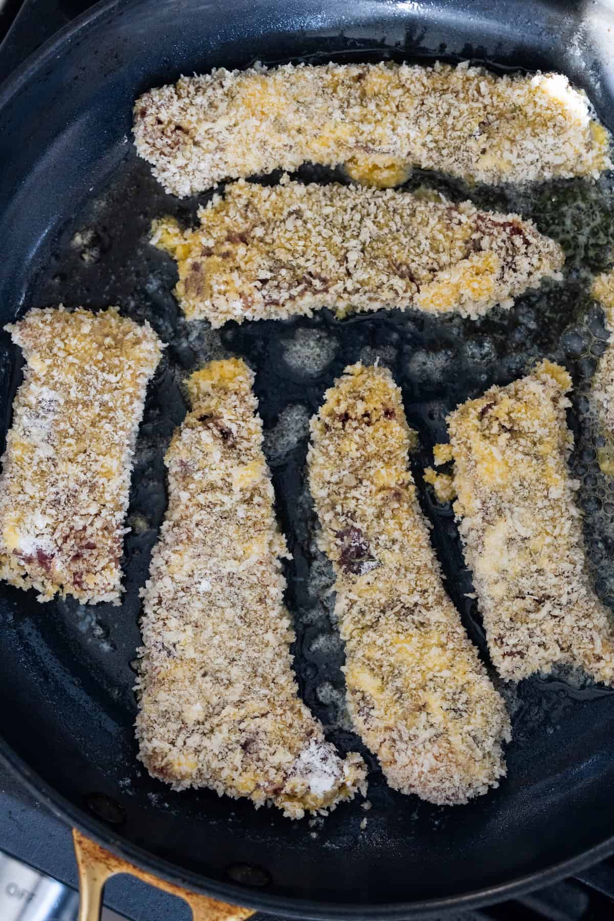 Breaded strips of meat, perfect for a classic Steak Fingers recipe, are being cooked in a black skillet with some oil visible on the surface.