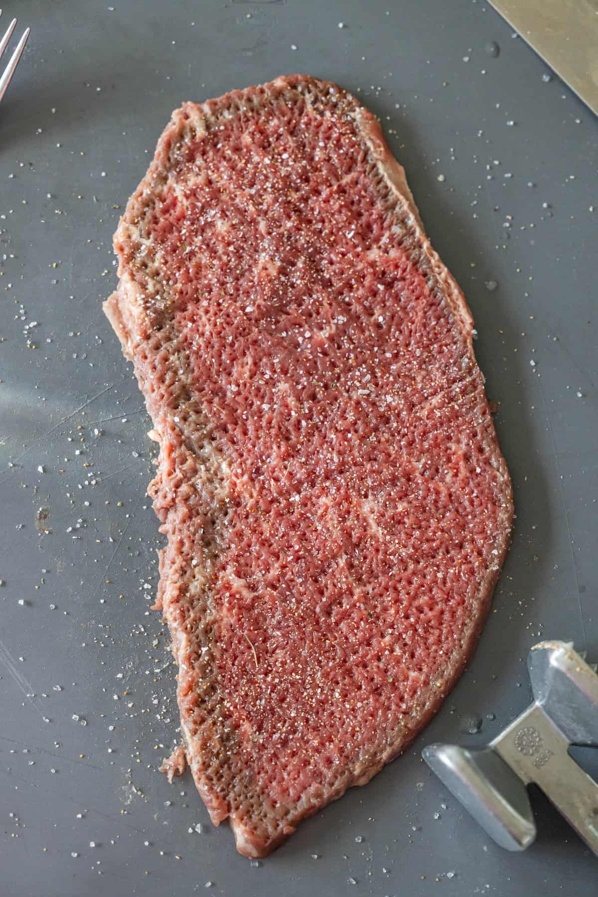 A raw piece of tenderized beef steak on a gray surface, sprinkled with salt and pepper, perfect for starting your Steak Fingers Recipe, with a meat mallet and fork nearby.