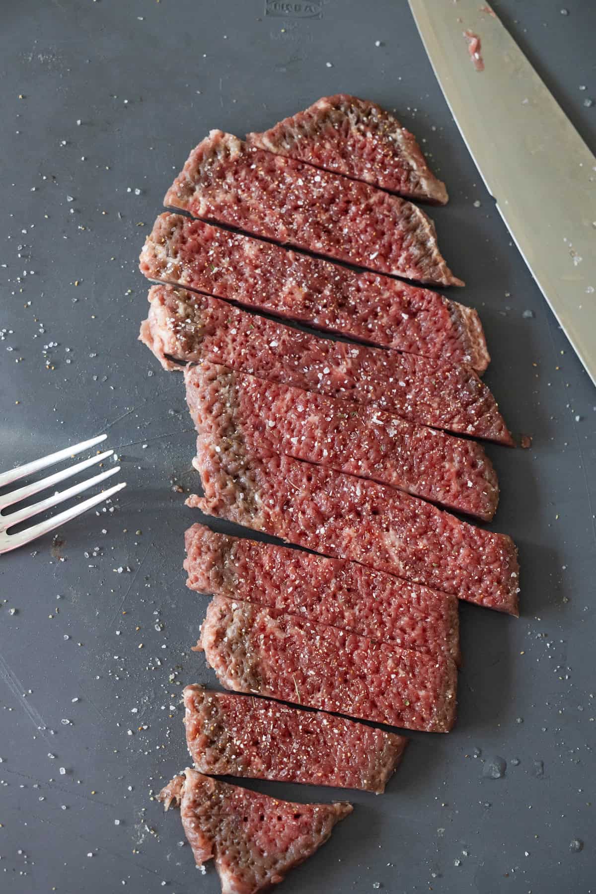 Slices of raw, seasoned beef on a gray cutting board with a fork and a knife nearby, ready for your favorite Steak Fingers recipe.