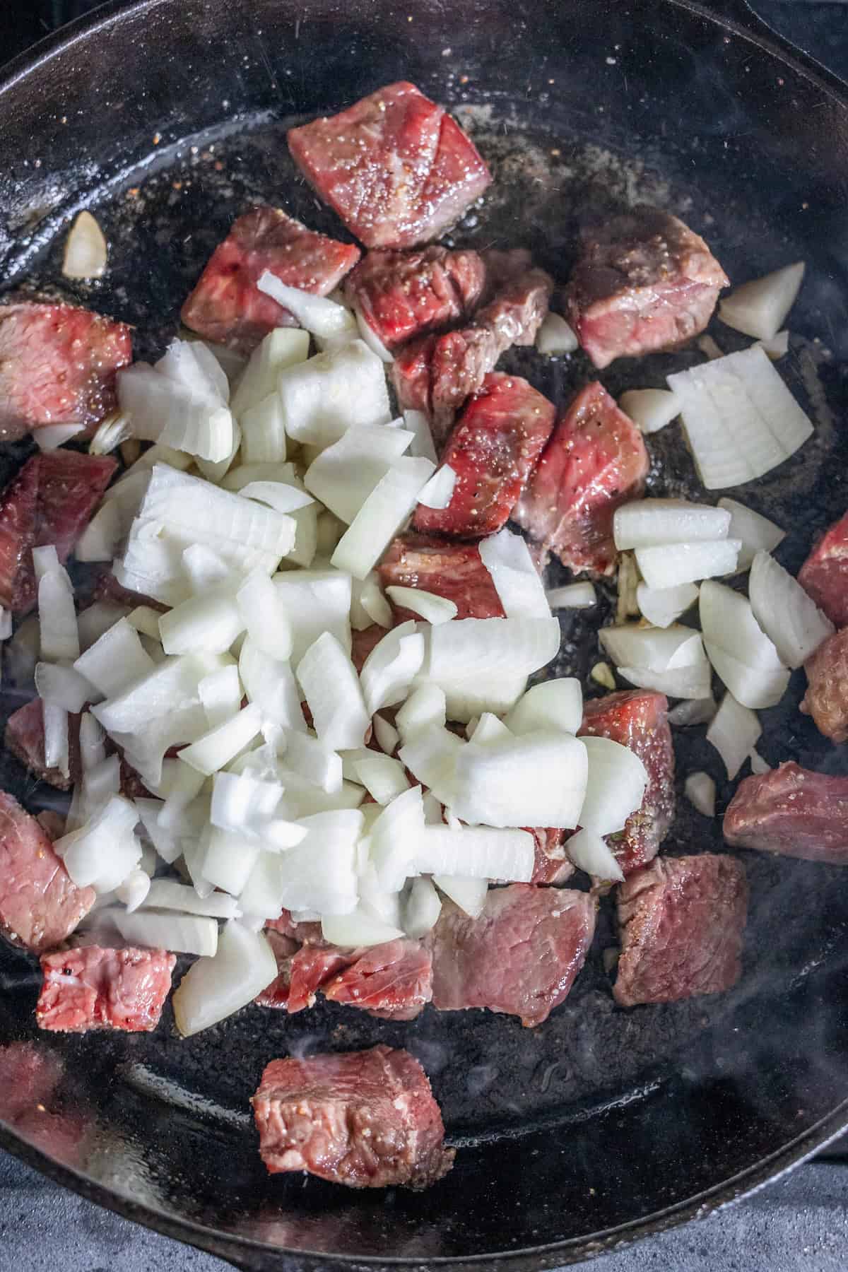 Cubed sirloin steak tips and chopped onions are being pan seared together in a black skillet.