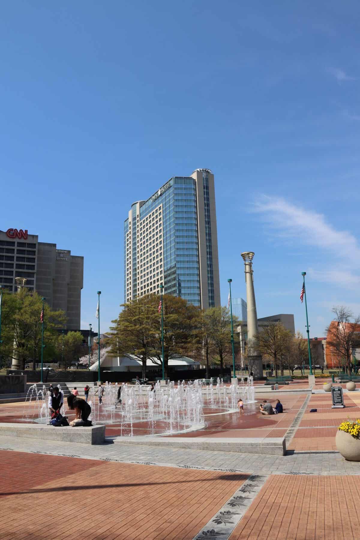 A city plaza with a central fountain, people sitting nearby, tall buildings in the background, and a clear blue sky overhead—a perfect scene featured in any Atlanta travel guide.