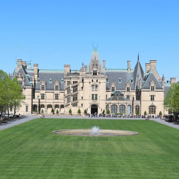 Large historic mansion with a steeply pitched roof, multiple chimneys, and ornate detailing reminiscent of the Biltmore estate, set behind a circular fountain and expansive lawn under a clear blue sky.