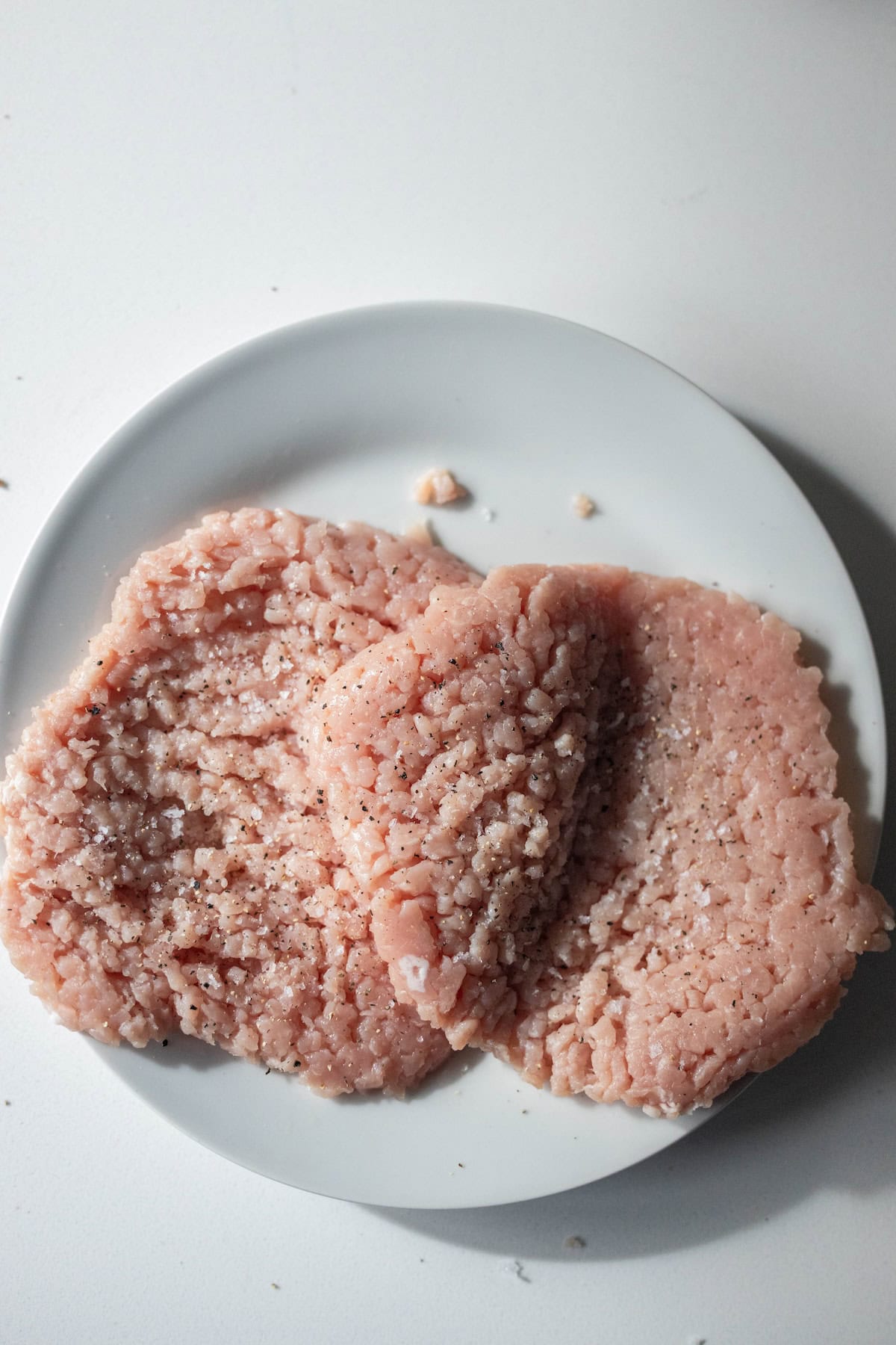 Three raw ground meat patties, perfect for making Pork Swiss Steak, with visible seasoning on a white plate, placed on a white surface.