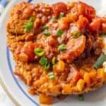A breaded meat patty topped with tomato-based sauce, diced vegetables, and chopped green onions on a plate.