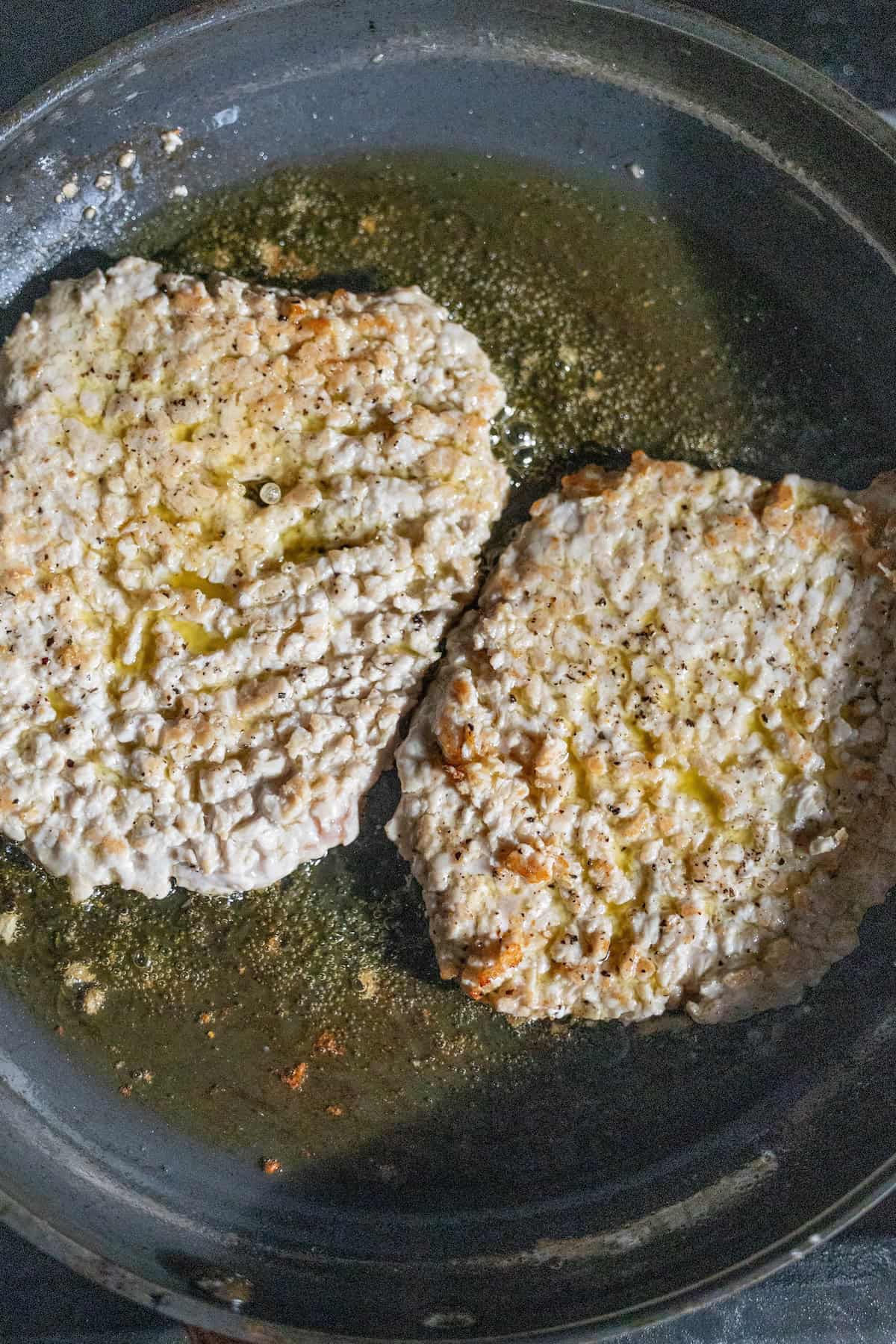 Two breaded and seasoned pieces of food, reminiscent of Pork Swiss Steak, are frying in oil in a black skillet.