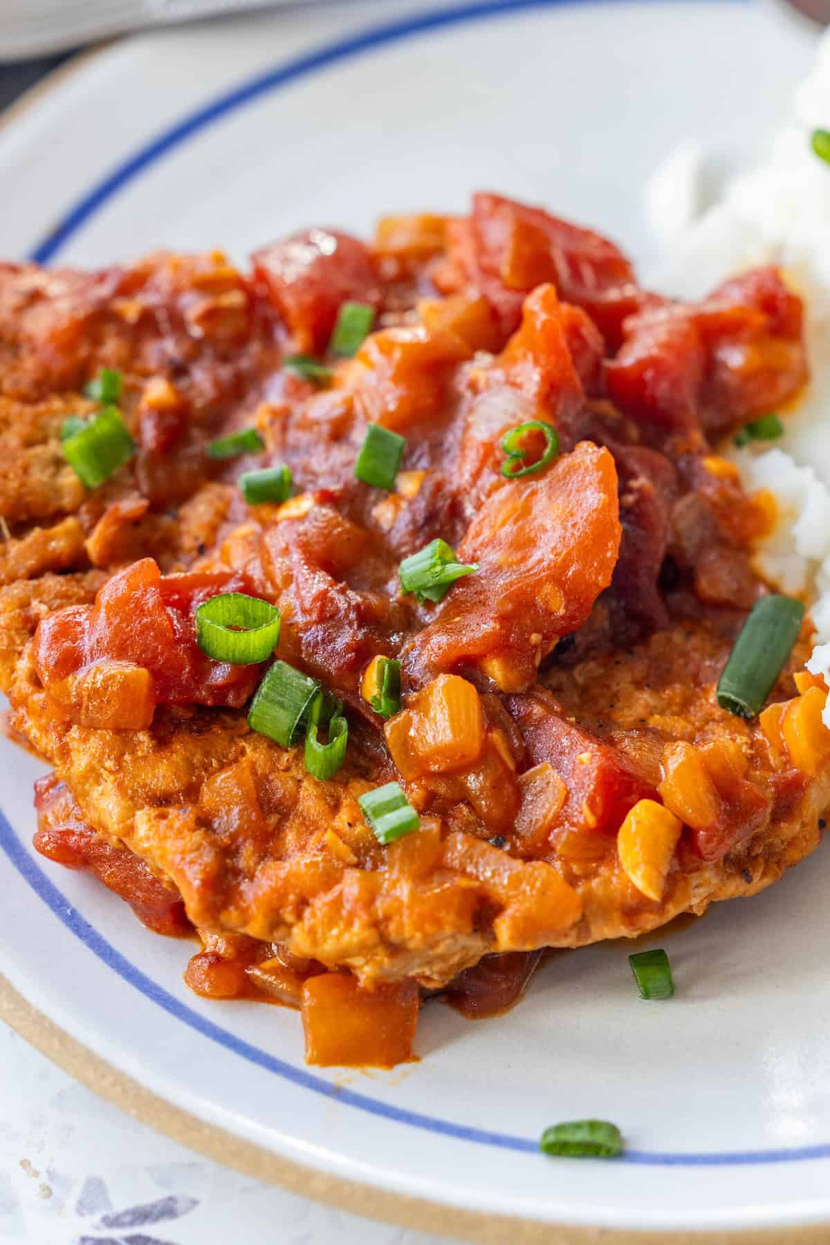 A plate featuring Pork Swiss Steak—a breaded meat cutlet topped with tomato-based sauce and chopped green onions—served alongside a portion of white rice.