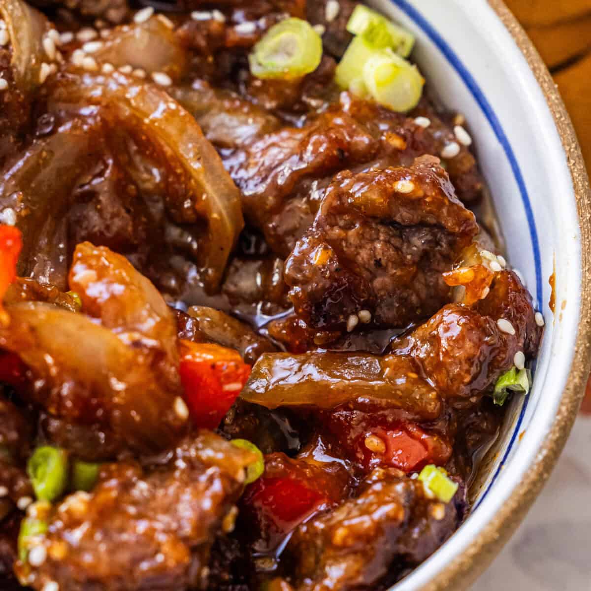 A close-up of better than takeout beef stir fry with onions, red bell peppers, green onions, and sesame seeds in a white bowl with a blue rim.
