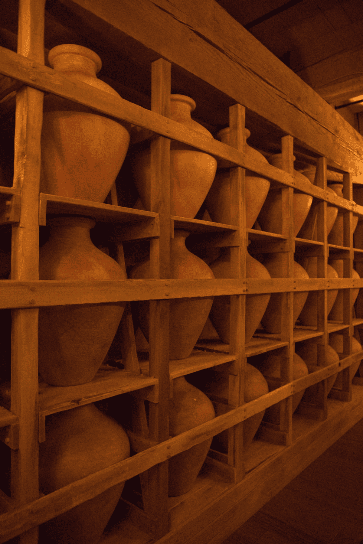 Large clay jars are stored upright in a wooden shelving unit with multiple compartments in a dimly lit room.