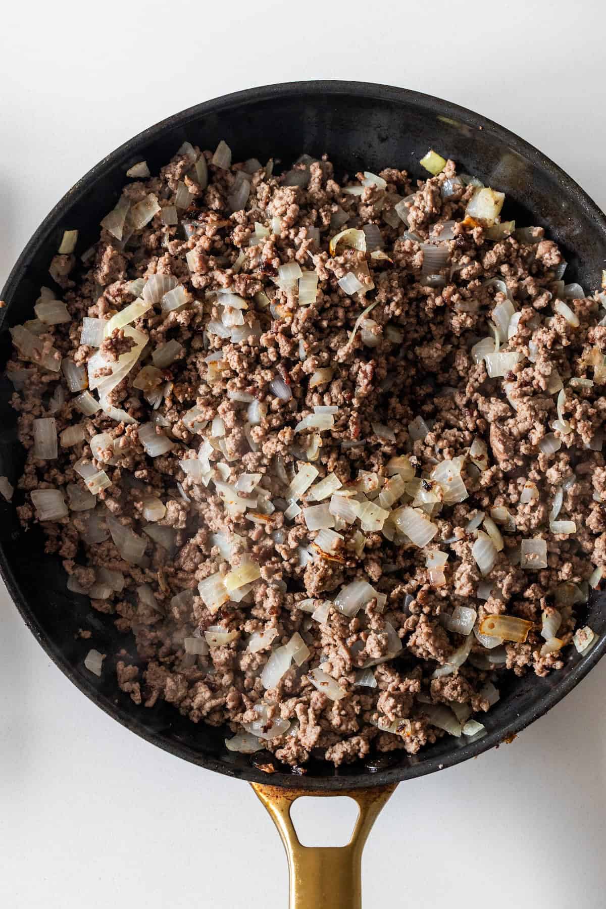 Ground beef and chopped onions cooking in a black skillet with a gold handle, viewed from above—perfect for starting your Dirty Rice Recipe or Baked Dirty Rice.