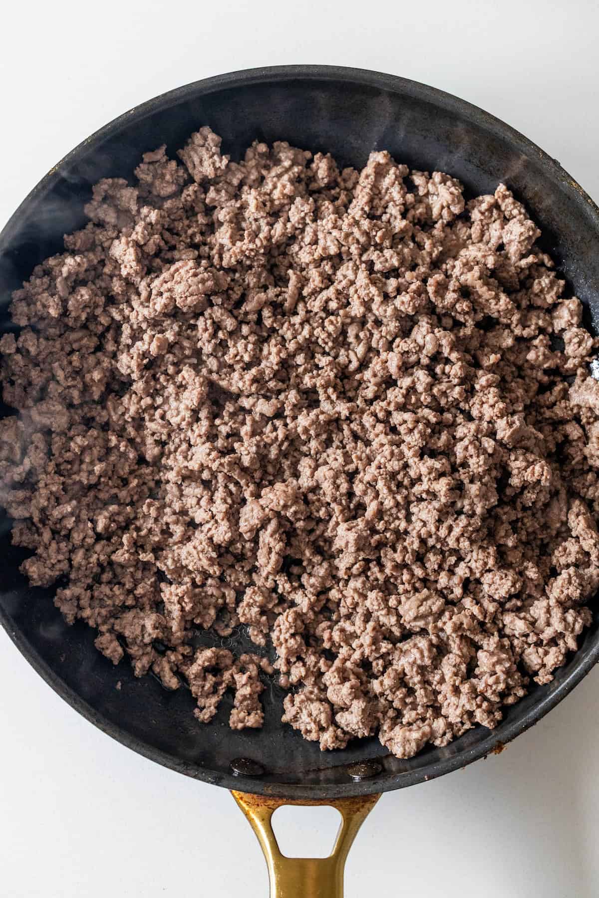 Cooked ground beef in a black skillet with a gold handle, viewed from above on a light surface—perfect for starting your next Dirty Rice Recipe.