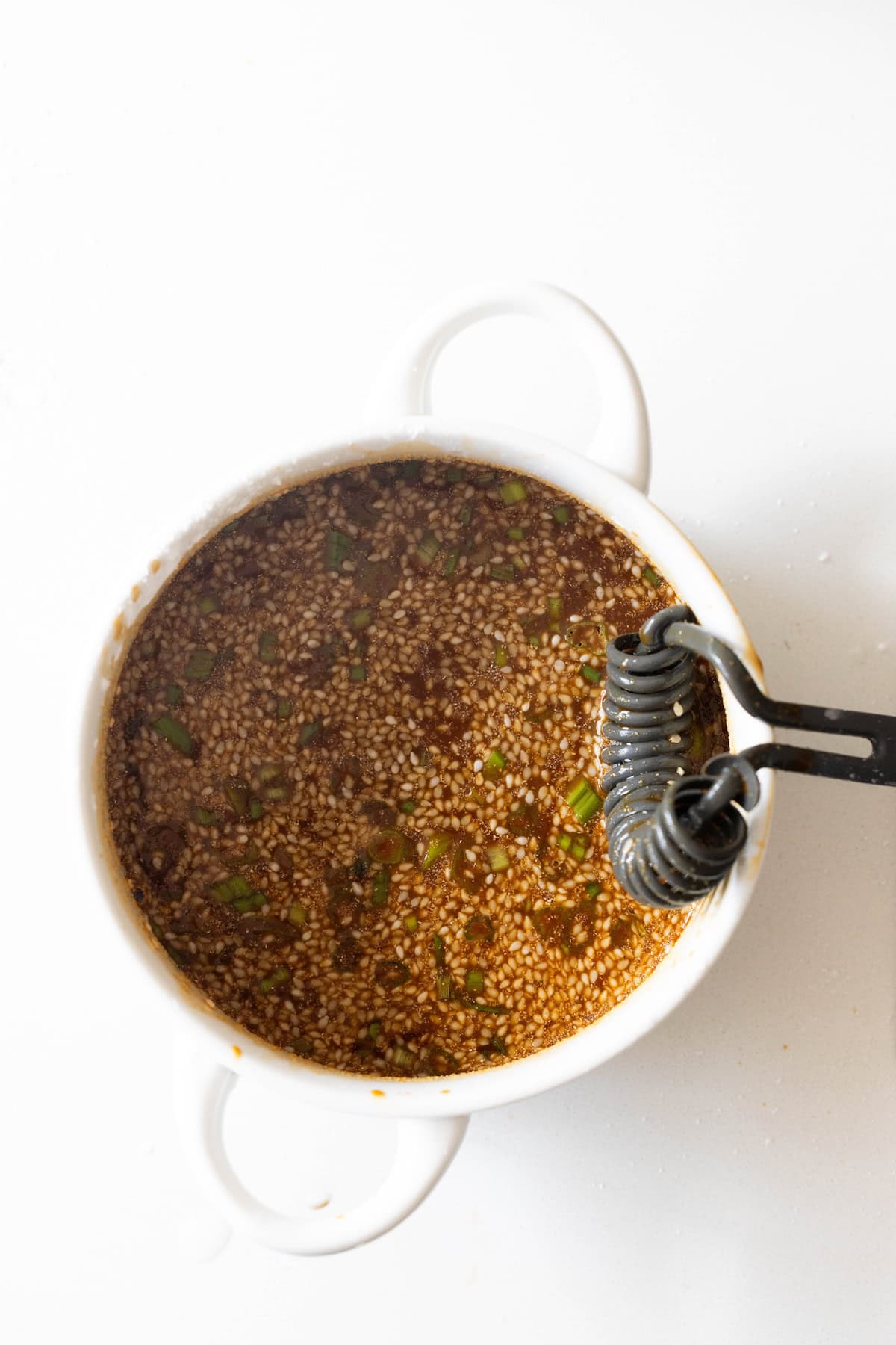 A small white bowl filled with a brown sesame seed sauce mixture, perfect for drizzling over pepper and beef stir fry, with chopped green herbs and a metal whisk resting inside.