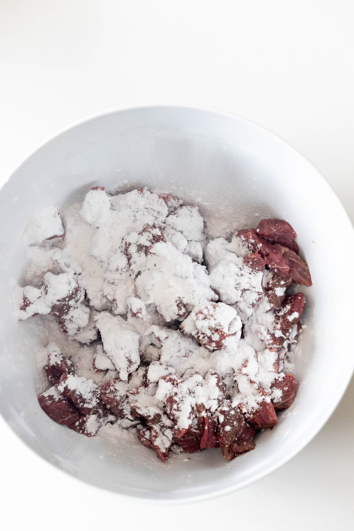 Chunks of raw meat in a white bowl are coated with a layer of white flour or starch, ready to become a delicious pepper and beef stir fry that's better than takeout.