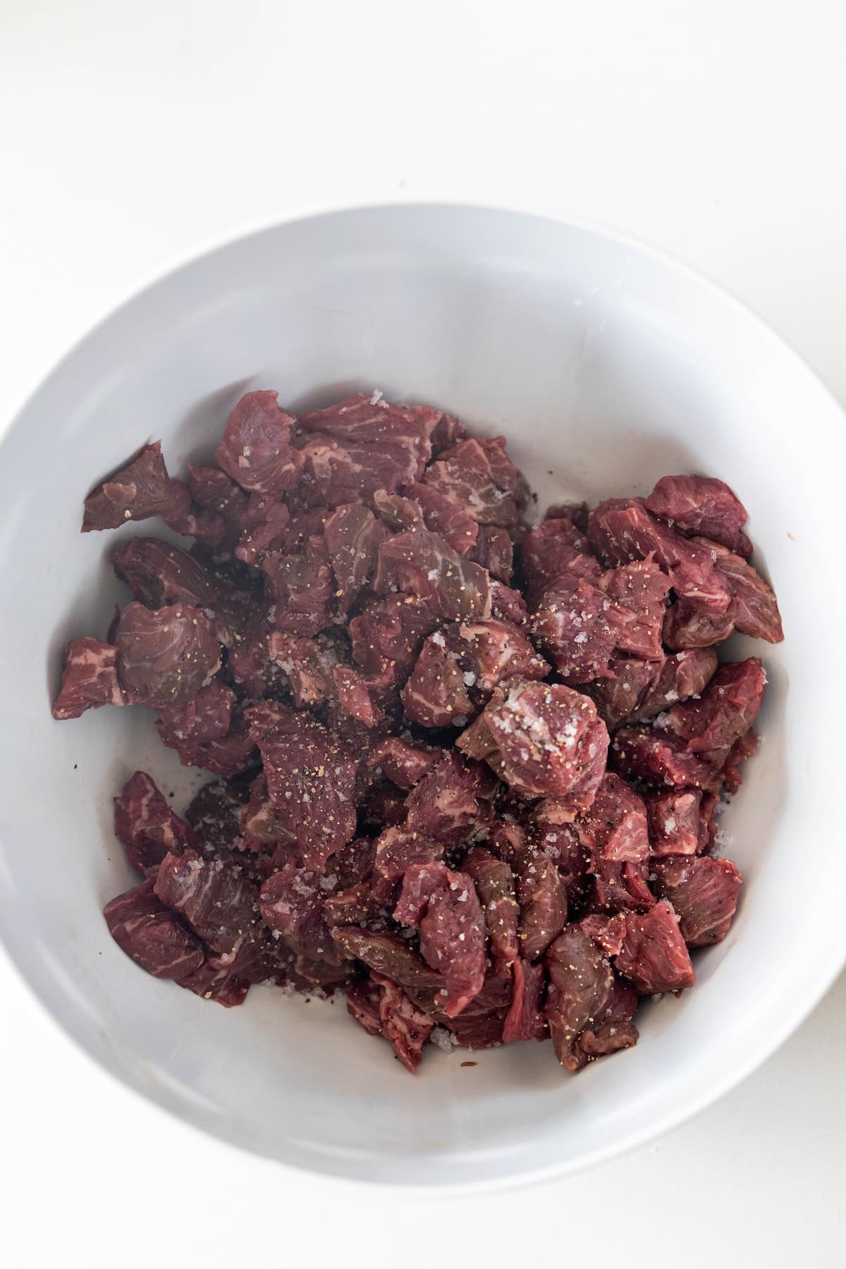 A white bowl containing raw, cubed beef seasoned with salt and pepper on a white surface—perfect for making a delicious pepper and beef stir fry at home.