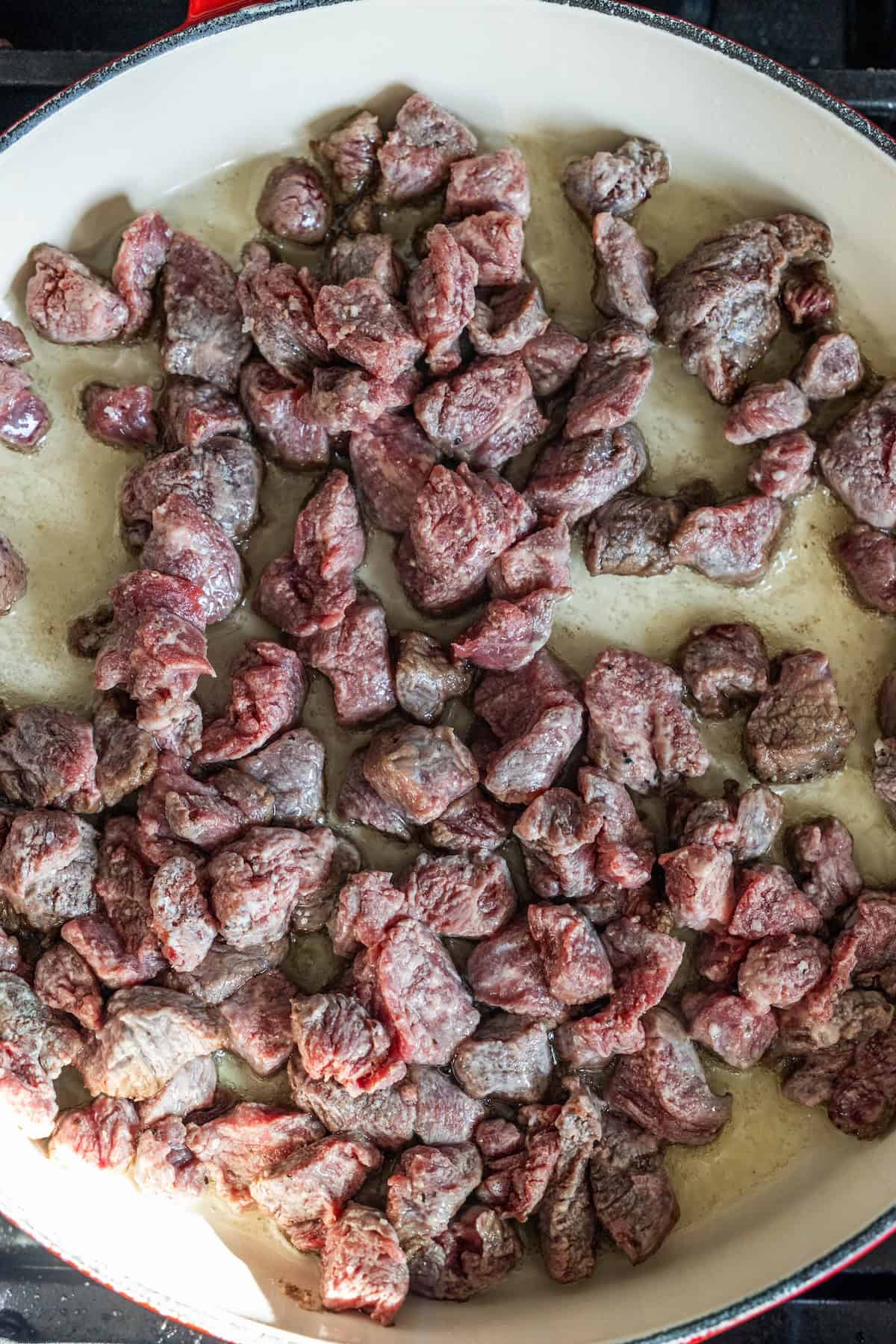 Chunks of raw beef browning in a large white skillet on a stovetop, perfect for a flavorful Stir Fry Peppers and Beef that's better than takeout.