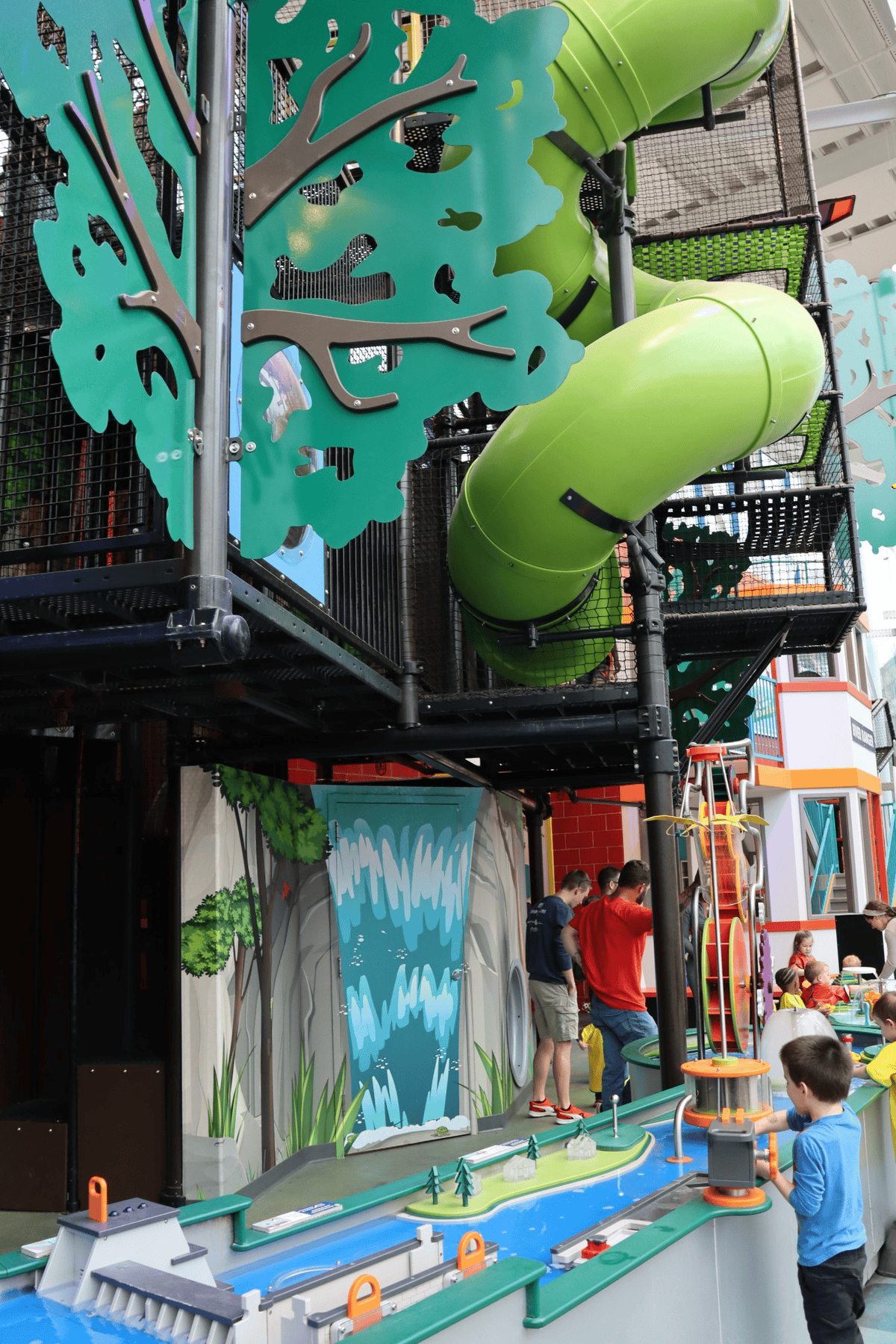 Children and adults play at an indoor activity center with green tube slides, climbing structures, and a water-themed play area.