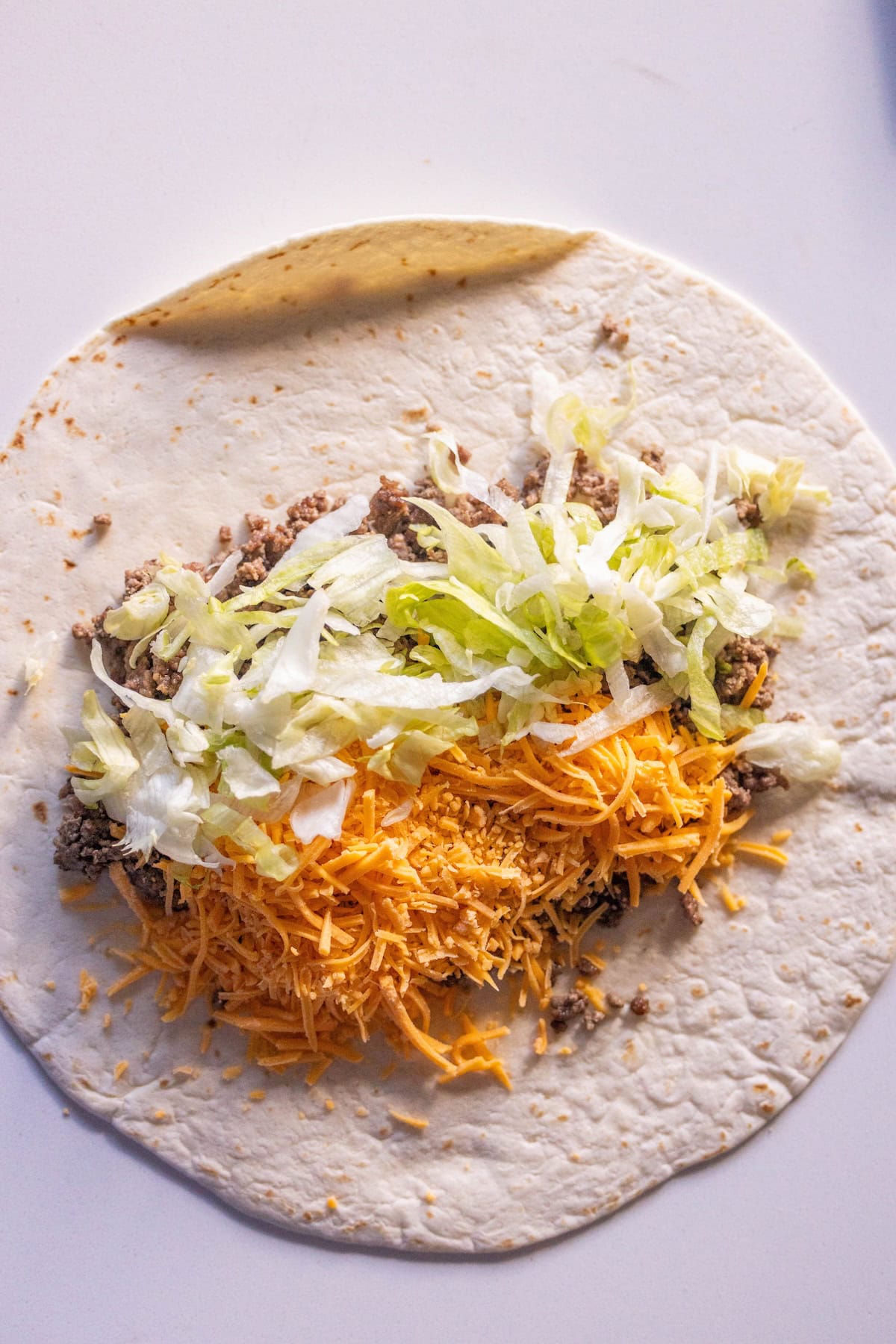 A flour tortilla topped with ground beef, shredded cheddar cheese, and lettuce on a white surface—these Easy Cheeseburger Wraps are ready to be wrapped and enjoyed.