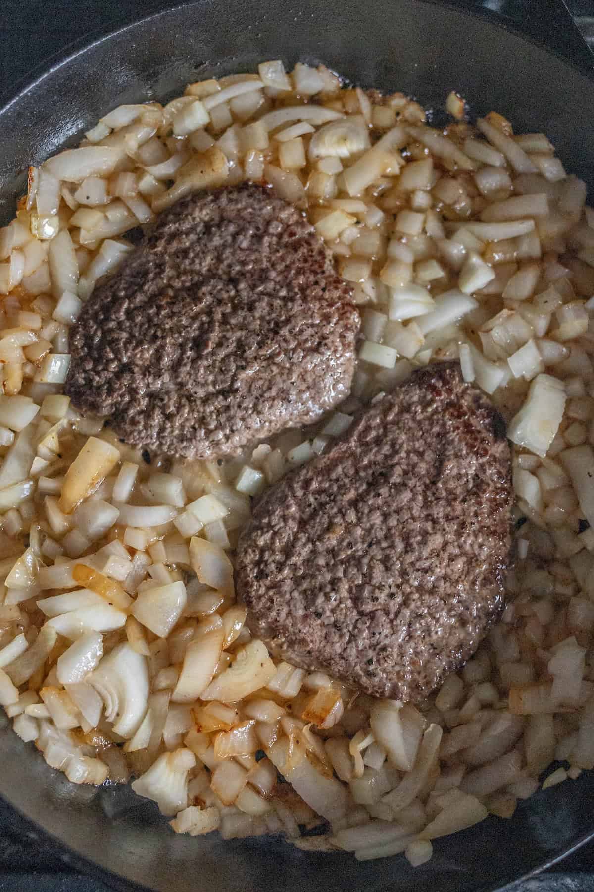 Two beef patties cooking in a skillet, surrounded by diced and sautéed onions—a classic country steak recipe in the making.
