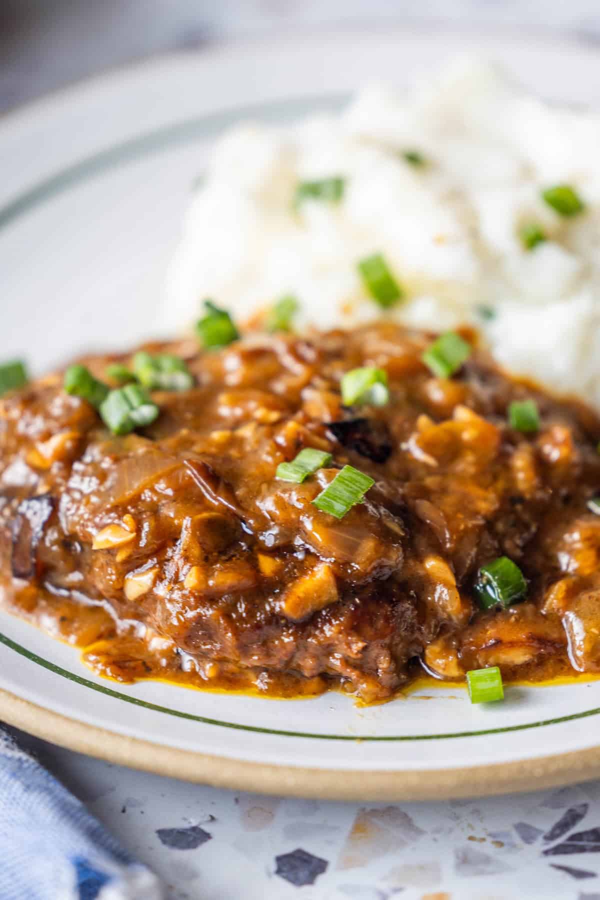 A plate with Salisbury steak, inspired by a classic Country Steak Recipe, topped with brown onion gravy, garnished with chopped green onions, and served alongside creamy mashed potatoes.