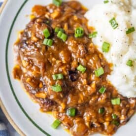 A plate with Salisbury steak, made using a classic country steak recipe, topped with brown onion gravy and served alongside mashed potatoes garnished with chopped green onions and black pepper.