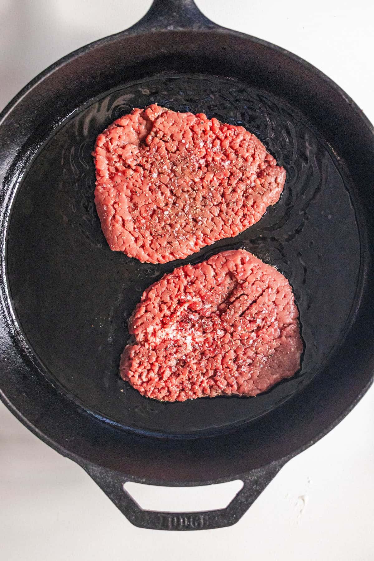 Two raw ground beef patties with salt and pepper are cooking in a black cast iron skillet with oil, perfect for starting a classic Country Steak Recipe.