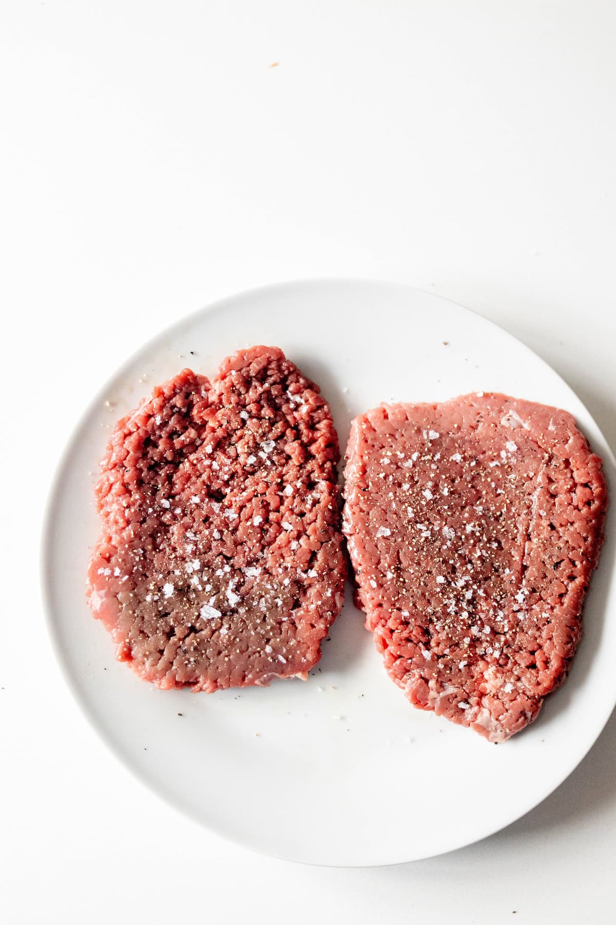 Two uncooked, seasoned beef patties on a white plate, sprinkled with salt and pepper, set against a white background—perfect for your next Country Steak Recipe.