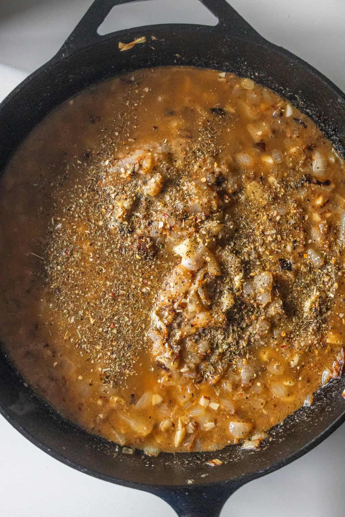 A cast iron skillet filled with a simmering mixture of chopped onions, broth, and various dried herbs and spices—a perfect base for your favorite country steak or classic gravy recipe.