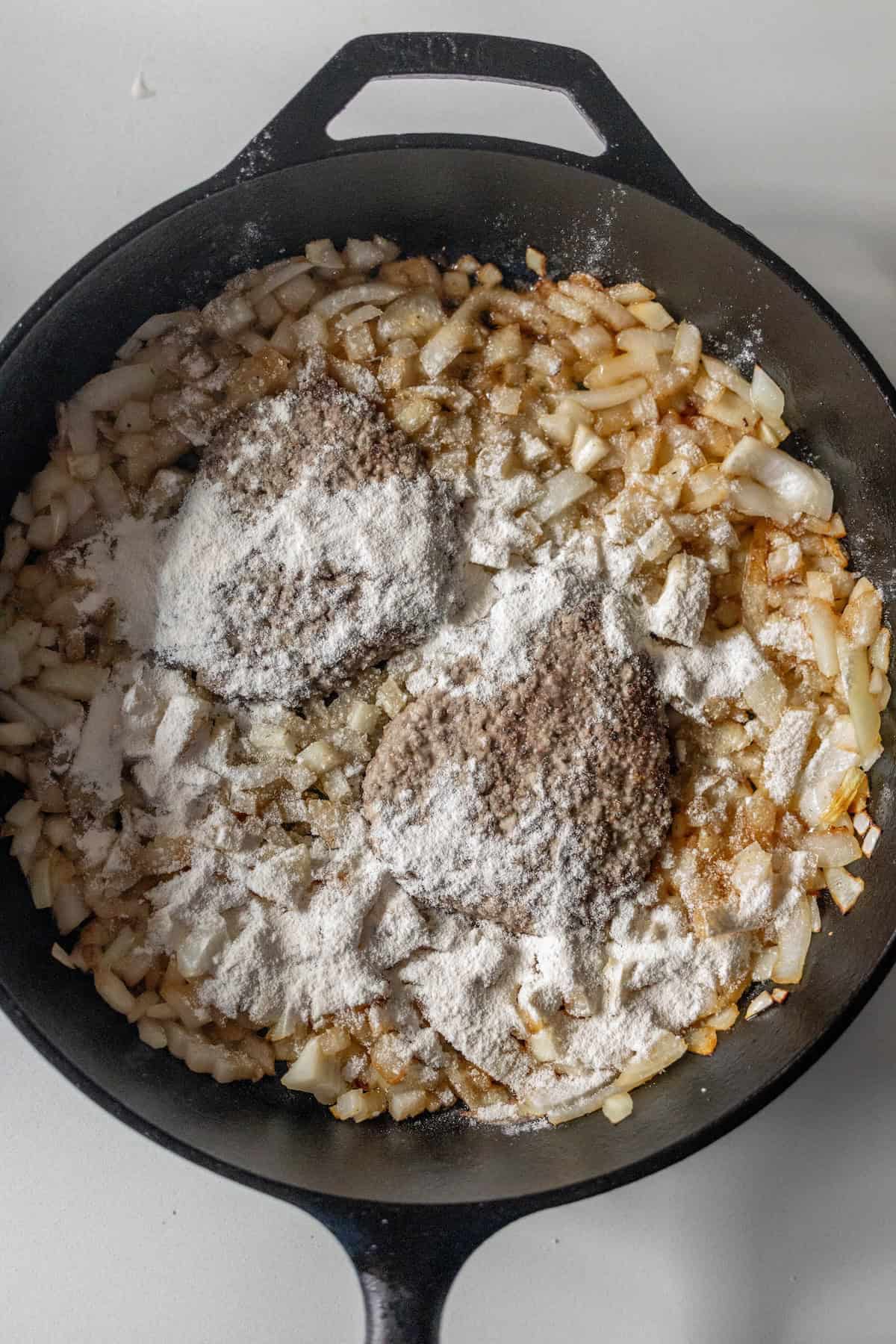 Chopped onions sautéing in a black cast iron pan with flour and two mounds of ground spices added on top—perfect beginnings for a classic Country Steak gravy recipe.