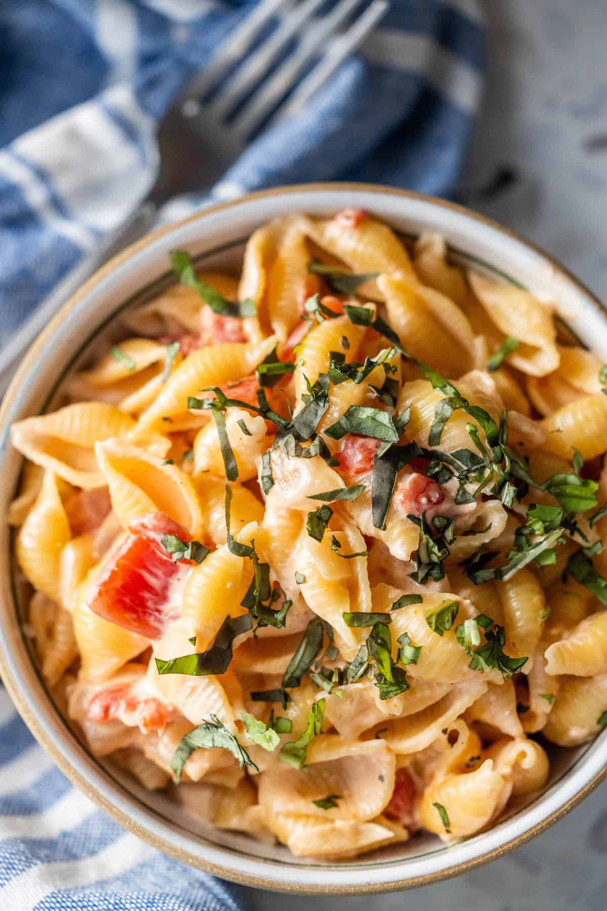 A bowl of shell pasta with creamy sauce, diced tomatoes, and chopped fresh basil, placed next to a blue and white cloth napkin and a fork.
