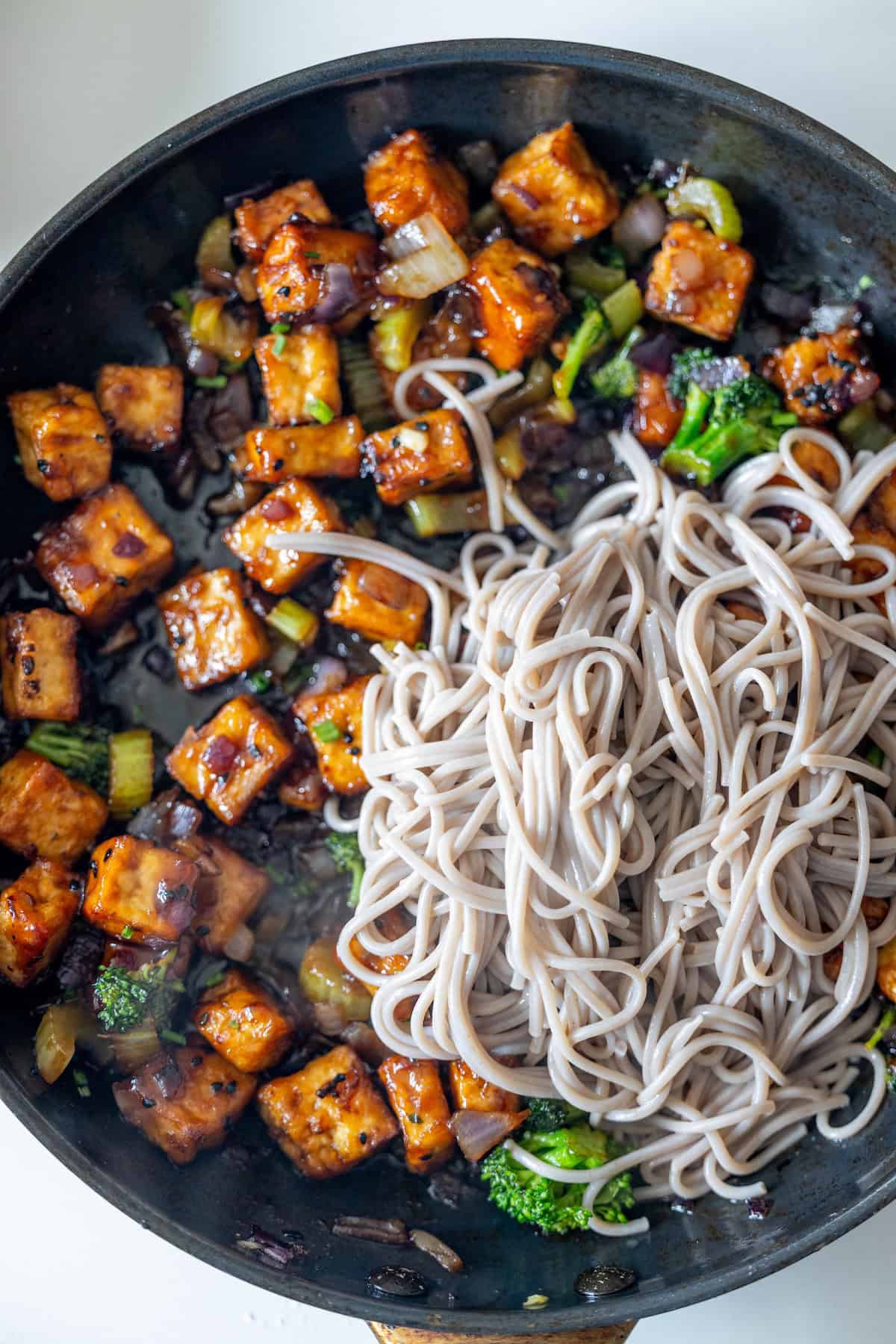 A skillet containing stir-fried vegetables and crispy tofu, with a portion of cooked soba noodles placed on one side.