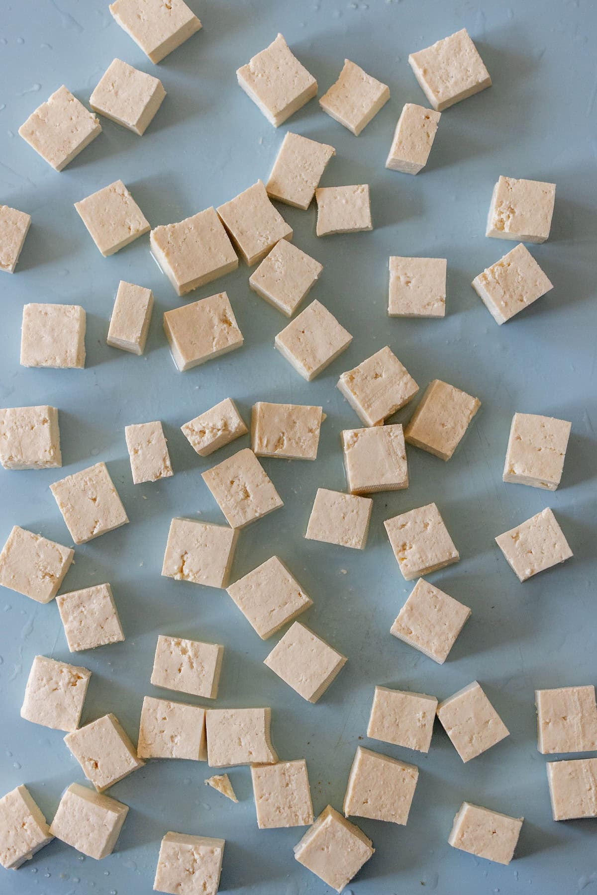 Cubed pieces of raw tofu are scattered on a light blue surface, ready to be turned into crispy tofu or added to a vibrant tofu stir fry.
