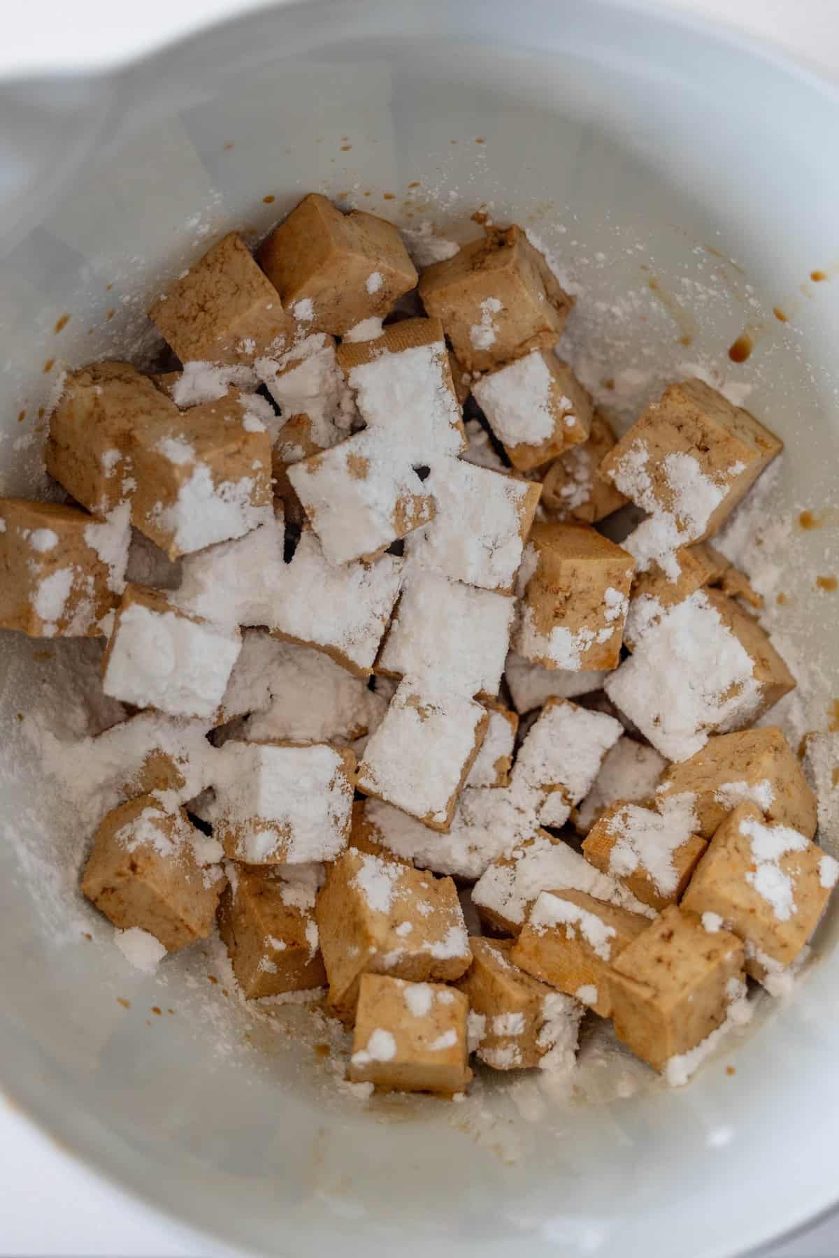 Crispy tofu cubes coated in a dark sauce and sprinkled with white powder, likely cornstarch, served in a white mixing bowl.