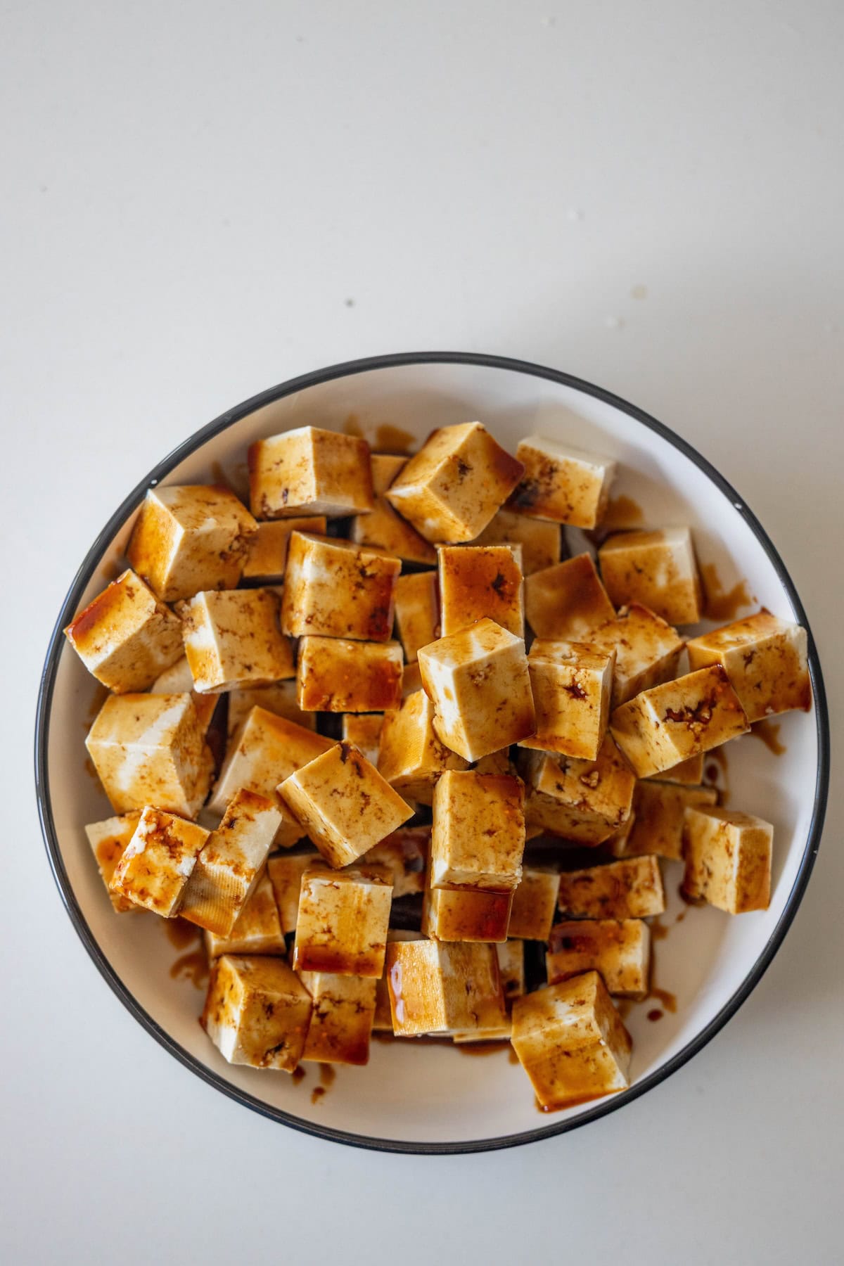 A bowl filled with evenly cut crispy tofu cubes coated in a dark marinade, perfect for adding to your favorite stir fry, placed on a white surface.