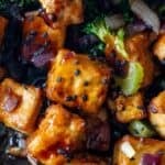 Close-up of crispy tofu cubes glazed to perfection, sprinkled with black sesame seeds, red onions, and broccoli pieces—an irresistible tofu stir fry.