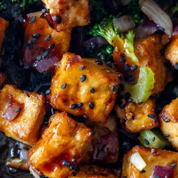Close-up of crispy tofu cubes glazed to perfection, sprinkled with black sesame seeds, red onions, and broccoli pieces—an irresistible tofu stir fry.
