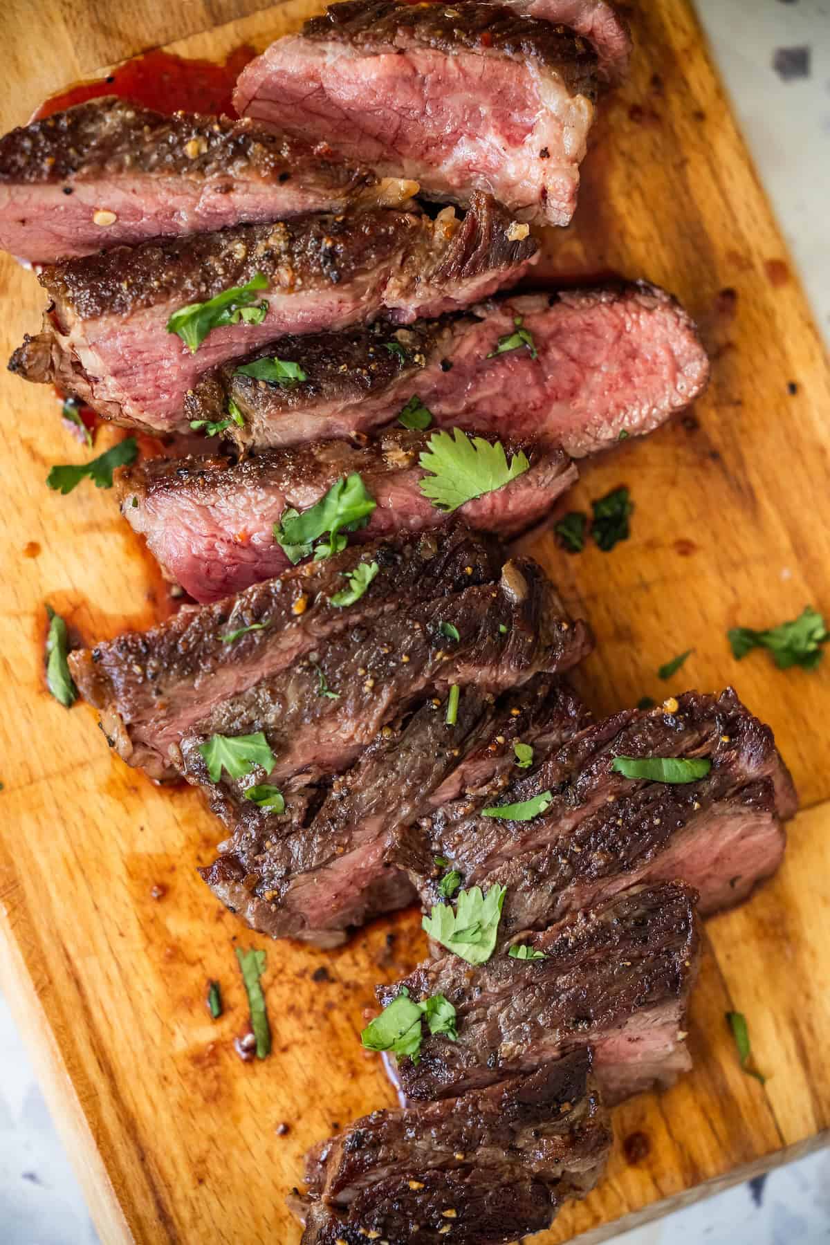Sliced medium-rare Denver Steak garnished with chopped herbs on a wooden cutting board.
