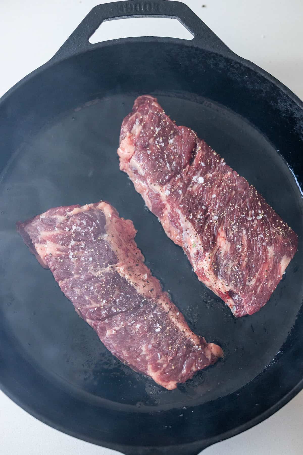 Two raw, seasoned Denver steak strips are placed in a black cast iron skillet, beginning to cook.