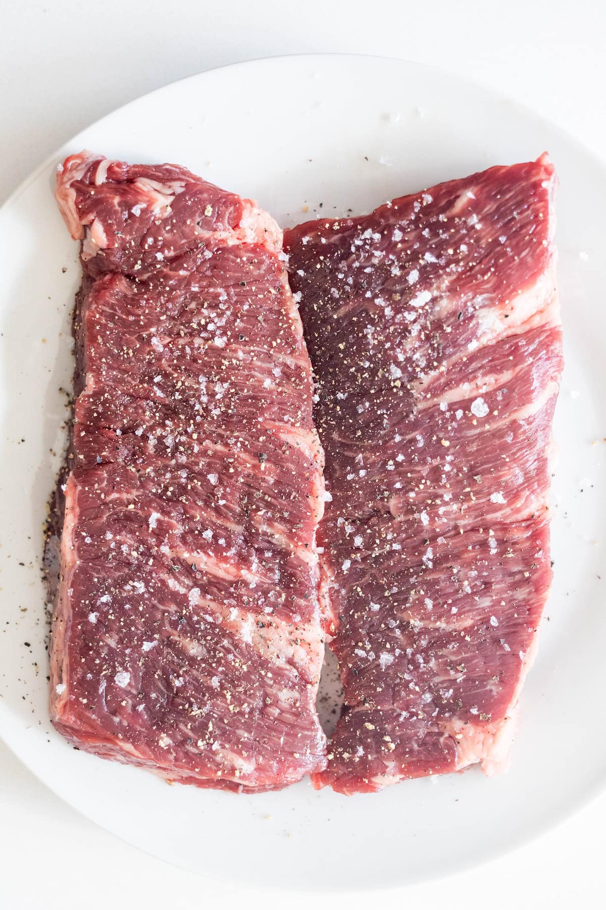 Two raw Denver Steak and skirt steak pieces on a white plate, sprinkled with coarse salt and ground black pepper.