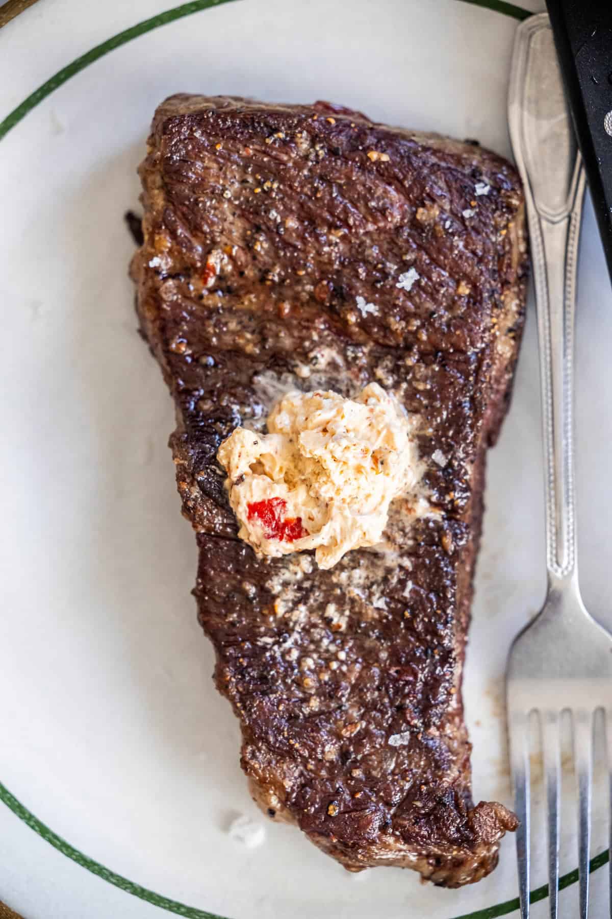 A cooked Denver Steak topped with a dollop of seasoned butter, served on a white plate with a green rim, next to a fork.