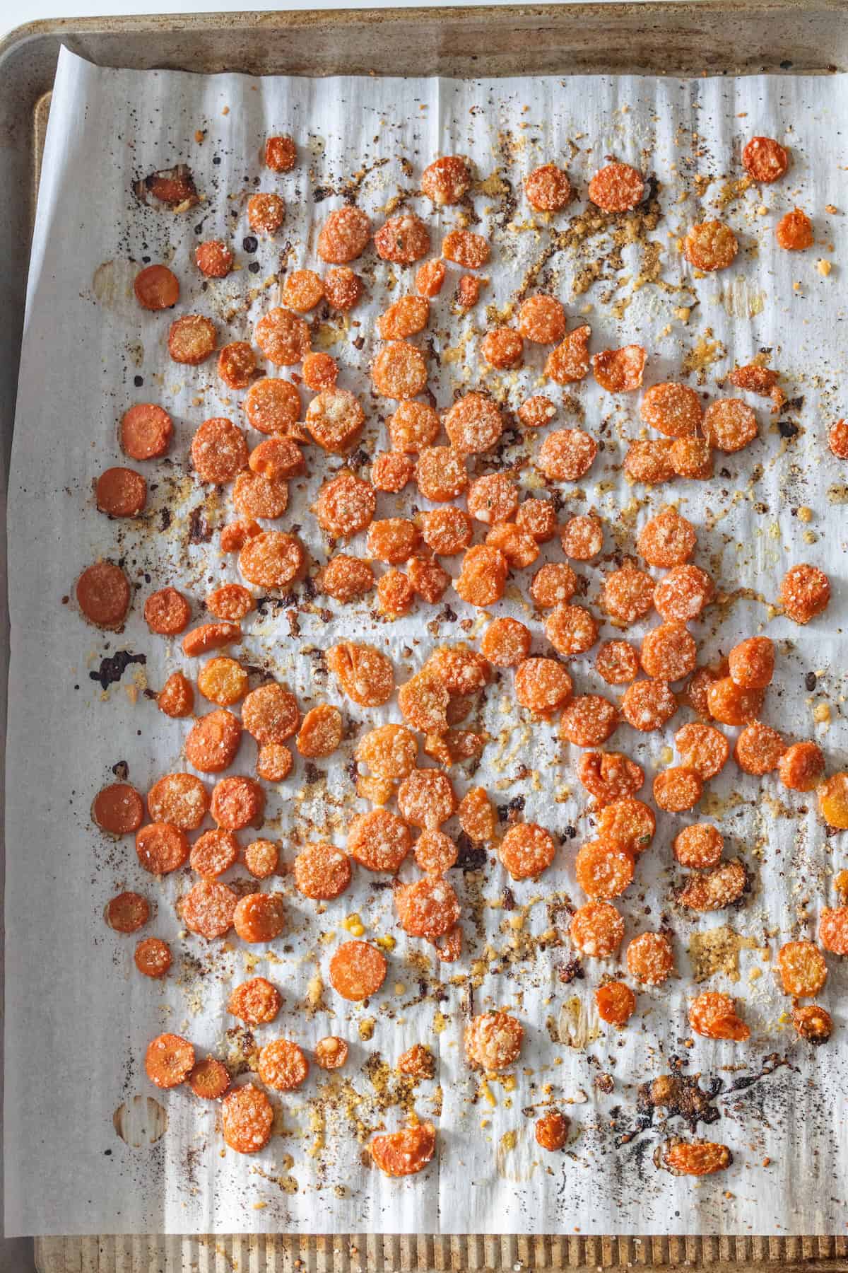 Baking sheet lined with parchment paper, topped with evenly spread Roasted Carrots slices with golden brown edges and some caramelized bits, perfect for making Garlic Parmesan flavor shine.