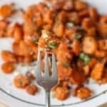 A fork holds a piece of roasted carrot garnished with herbs and grated cheese, with more roasted carrots blurred on a plate in the background.