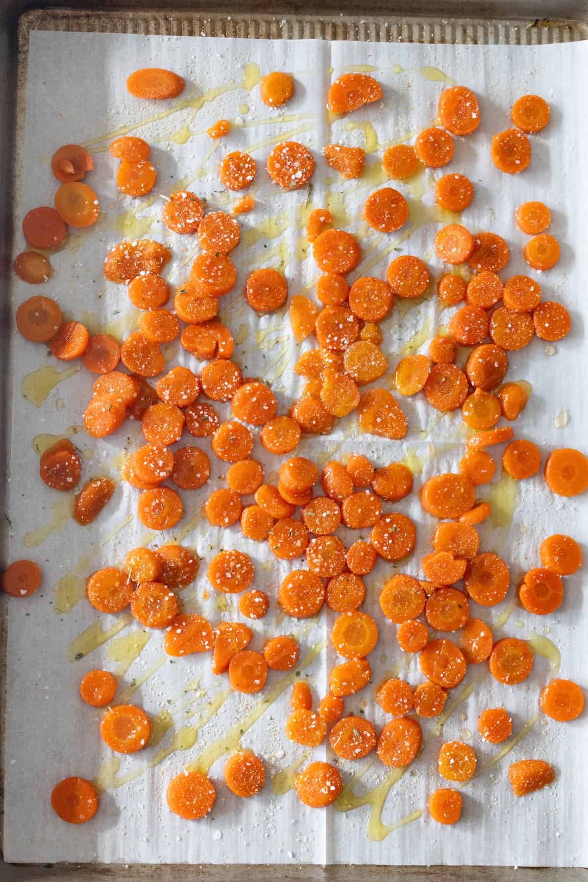 Sliced carrots spread on parchment paper, drizzled with oil and sprinkled with salt, pepper, and garlic Parmesan, ready for roasting on a baking sheet.