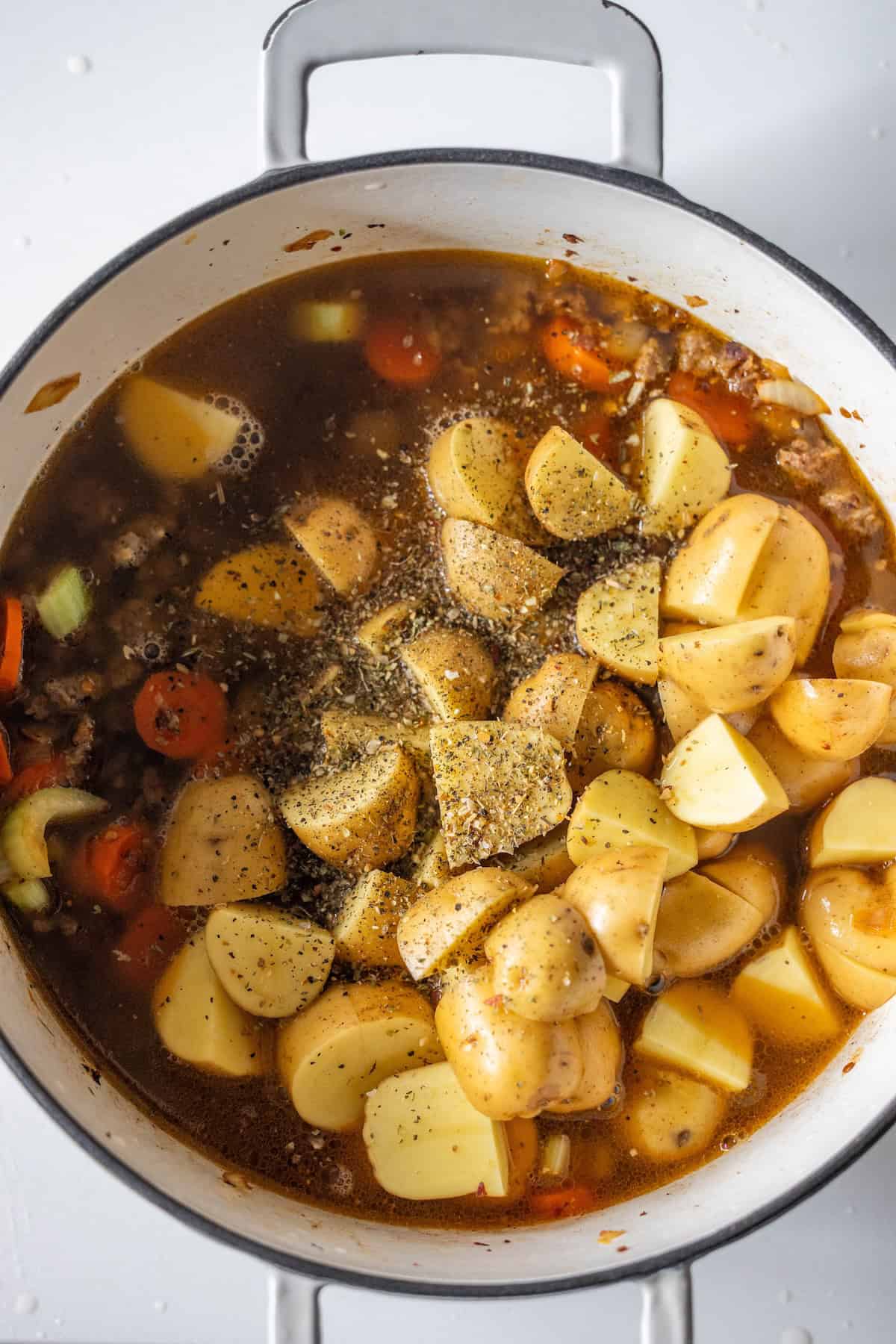 A pot filled with hearty Vegetable Soup—broth, chopped potatoes, carrots, celery, and ground sausage—topped with a sprinkle of black pepper and seasonings, sitting on a white surface.