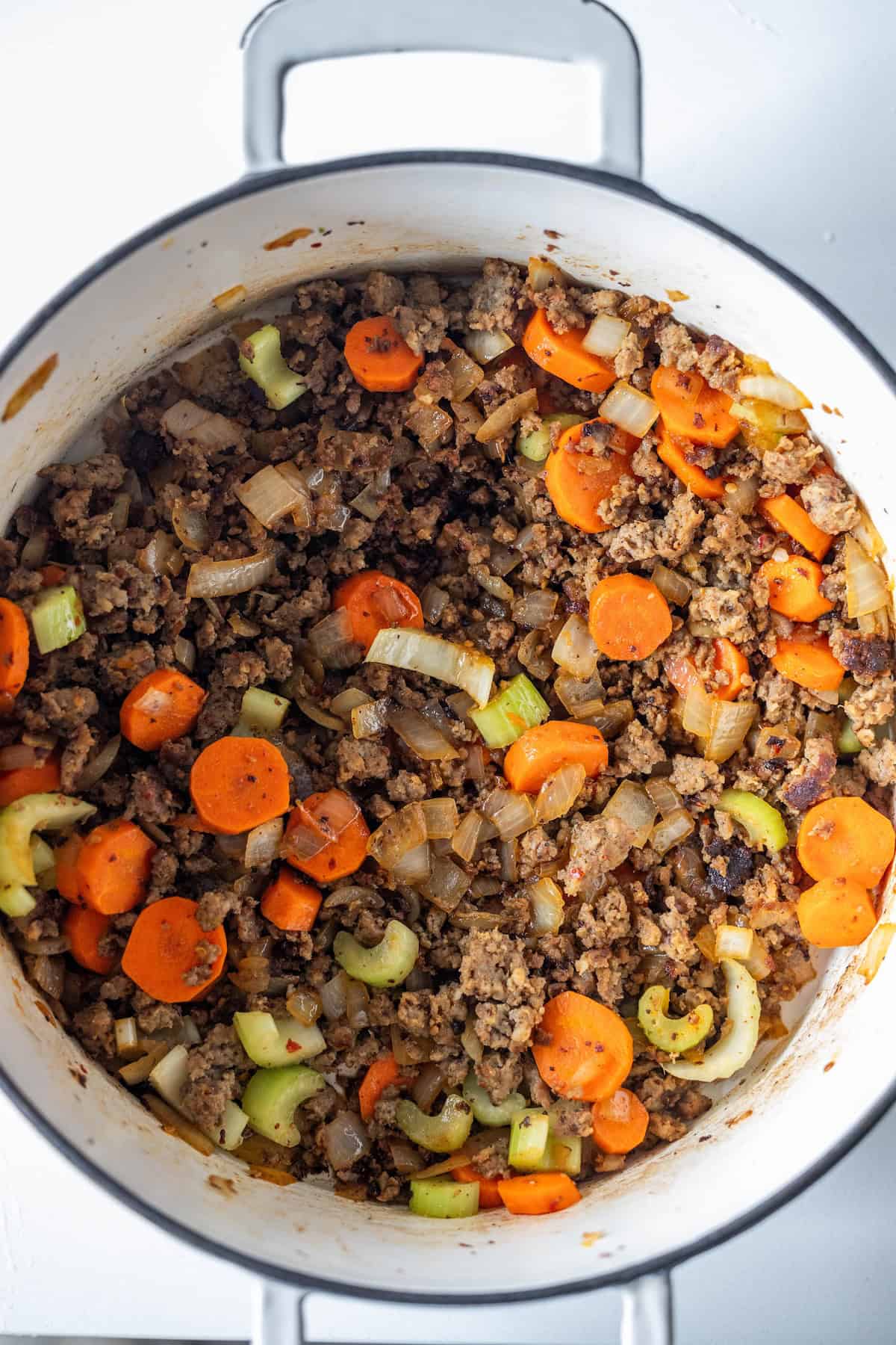 Ground meat, sausage, chopped carrots, celery, and onions simmer together in a white pot for a hearty vegetable soup.
