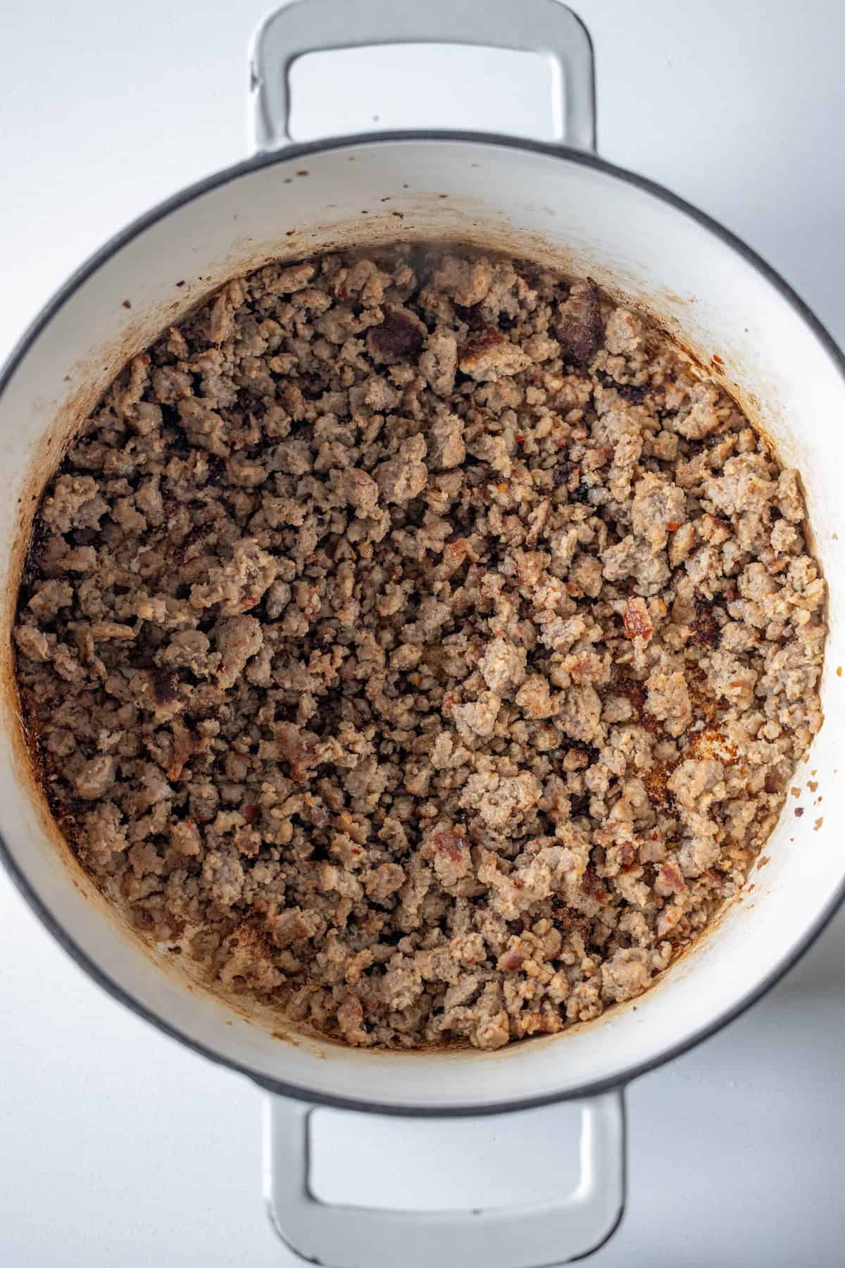 Cooked ground sausage fills a white pot viewed from above, with browned bits visible around the edges and some oil residue at the bottom—perfect for adding to a hearty vegetable soup.