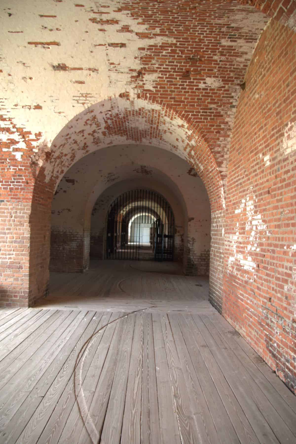 A view of arched brick doorways and wooden floors in an old fort or historic building, with a gated area visible in the background.