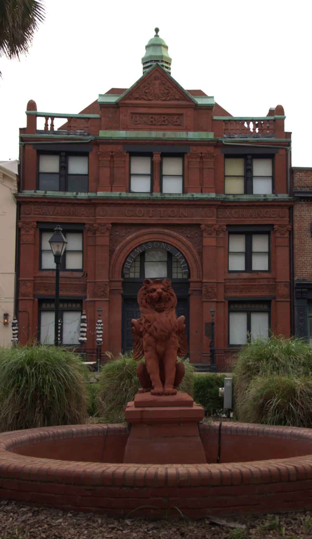 A red brick building labeled "Savannah Cotton Exchange" stands behind a red lion statue on a fountain, surrounded by greenery.