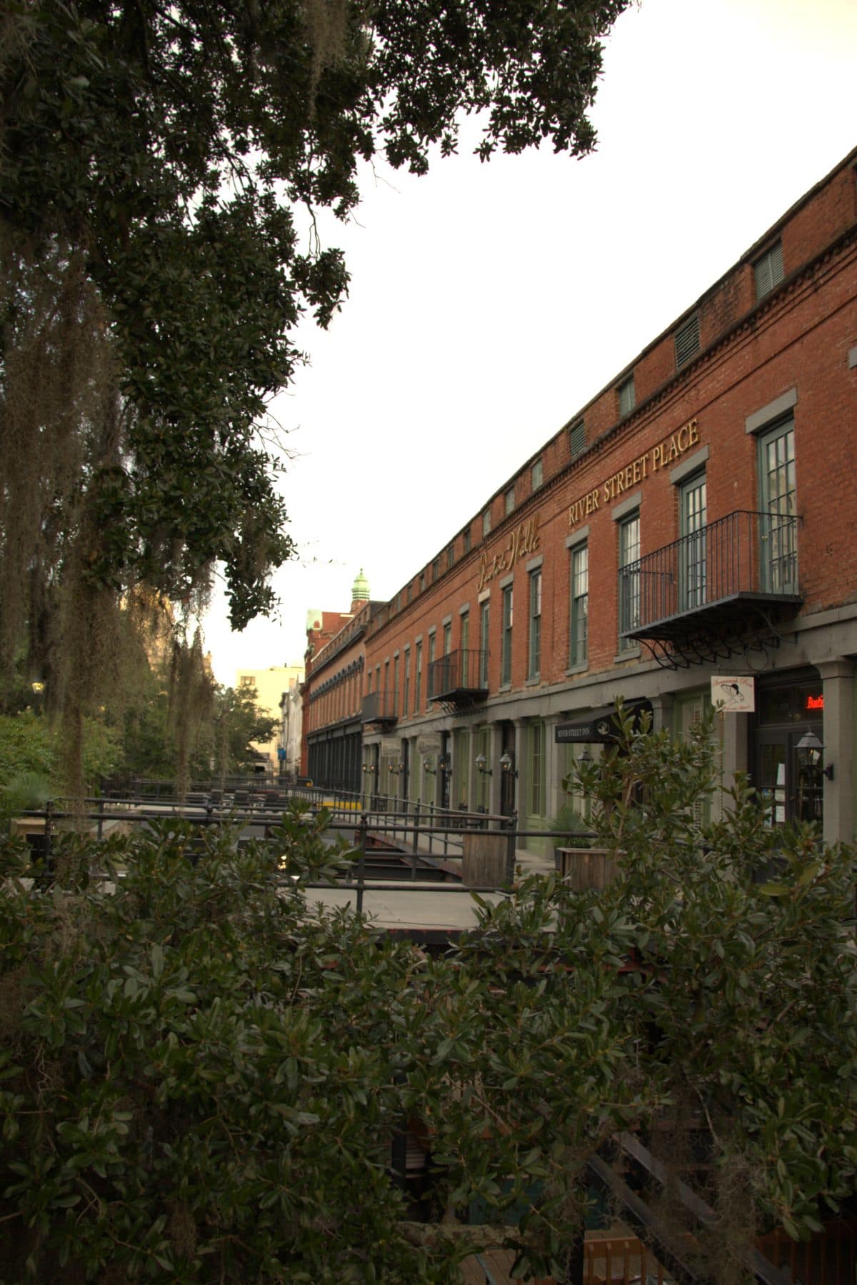 A row of historic red brick buildings labeled "River Street Place" with iron balconies, trees, and greenery in the foreground.